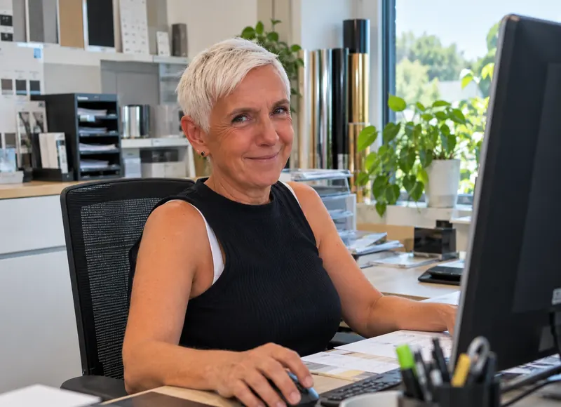 Silvia Nöll lächelt am Computer in ihrem hellen Büro bei Westerwald-Folien, im Hintergrund Folienmuster und Pflanzen am Fenster